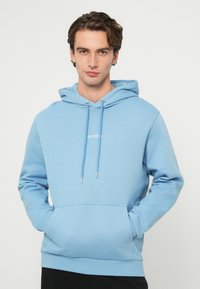 Fiatal barna hajú férfi, aki világoskék kapucnis pulóvert visel első zsebbel, és egy sima fehér háttér előtt áll.