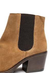 Bottine en suède marron avec un panneau latéral en élastique noir côtelé et un talon empilé, présentant des coutures visibles et une texture lisse.