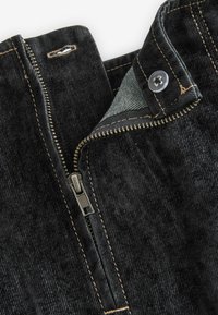 Tissu en denim noir avec fermeture éclair, couture visible et détail de bouton. Le col présente un doublure en tissu léger.