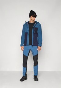 Plava flis jakna s tamnoplavim detaljima, uparena s plavo-crnim hlačama koje imaju trokutasti uzorak. Crna kapa i sunčane naočale upotpunjuju izgled.