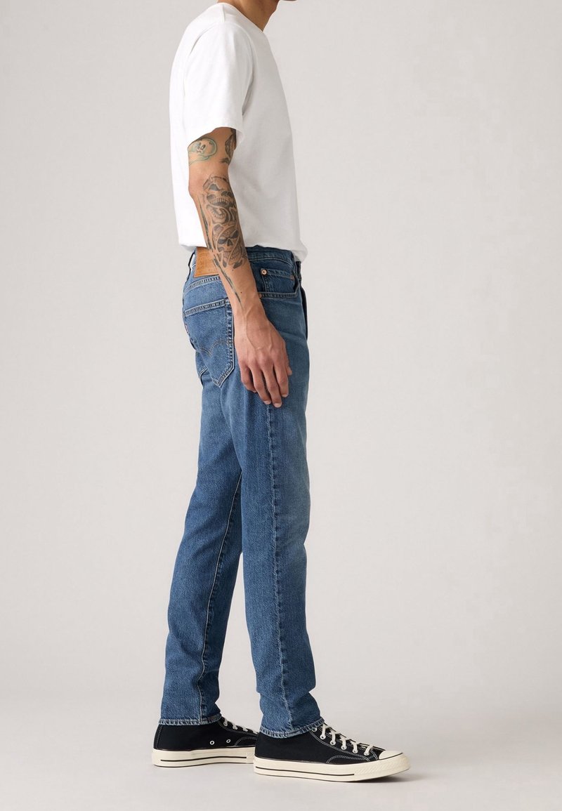 Homme portant un t-shirt blanc, un jean bleu, des baskets noires et des tatouages visibles sur le bras gauche, debout de profil sur un fond uni.