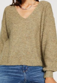 Maglione lavorato a maglia marrone-verde con scollo a V, maniche ampie e texture a coste. Abbinato a jeans in denim blu, mostra uno stile casual.