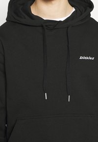 Detailní záběr osoby v černé mikině s kapucí, na které je vytištěno logo "Dickies" na hrudi, a se stáhnutými šňůrkami visícími dolů přes bílou košili.