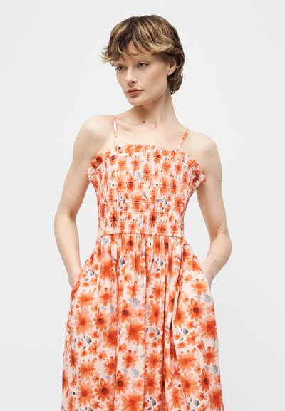 Γυναίκα με κοντά μαλλιά, ντυμένη με πορτοκαλί αμάνικο φόρεμα με floral σχέδιο και κορσέ με λάστιχο, στέκεται με τα χέρια στις τσέπες μπροστά σε απλό φόντο.