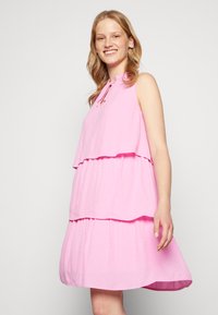 Vestido sem mangas de um tom rosa claro, com duas camadas, apresentando um tecido texturizado e um detalhe de laço na gola.