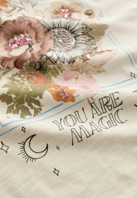 Tissu crème avec des imprimés floraux aux couleurs atténuées, parsemé de sequins et portant le texte "VOUS ÊTES MAGIQUE" en police noire en gras.