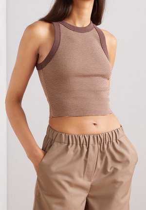 Nainen, joka pitää yllään hihattoman, uritetun ruskean lyhytpaitaisen ja beigejä korkeavyötäröisiä housuja, kädet taskuissa yksiväristä taustaa vasten.