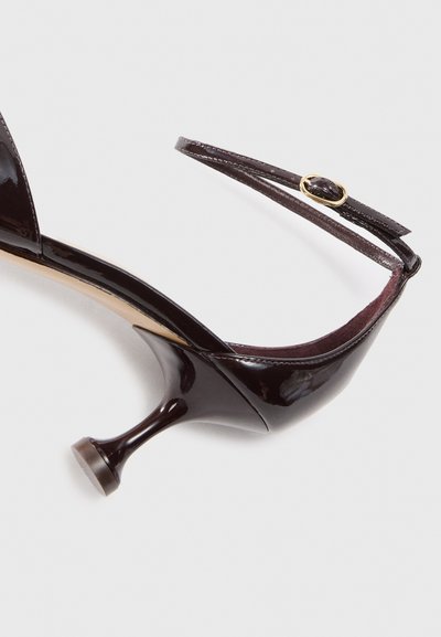 Black patent leather high-heeled shoe with a slender heel and adjustable ankle strap featuring a gold buckle. Rounded toe with smooth texture.