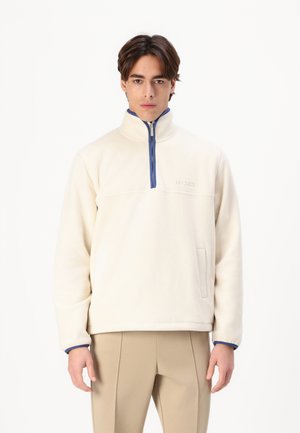 Cream-colored fleece pullover with a half-zip collar. It features a blue zipper, a front pocket, and a subtle logo on the chest.