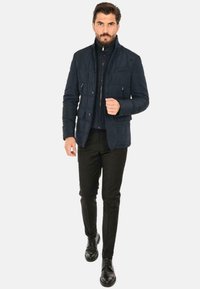 Giubbotto imbottito navy con colletto alto, multiple tasche e dettagli con zip. Abbinato a pantaloni neri slim-fit e scarpe nere.