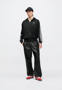 Čierna športová bunda s bielymi pruhmi, čierne nohavice a červené topánky. Bunda má zips a logo na hrudi.