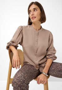 Camicia button-up color marrone chiaro con maniche a sbuffo, texture morbida e colletto rotondo. Abbinata a leggings scuri decorati con una stampa leopardata.