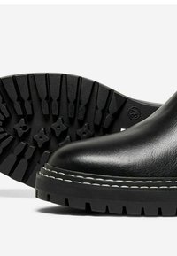 Bota de cuero negro con una suela gruesa y profundos surcos, y costuras blancas en contraste a lo largo del borde. Textura suave y puntera redondeada.