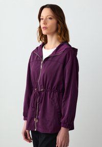 Paarse lichte windjack met een capuchon, trekkoord in de taille, ritsluiting en elastische manchetten. Glad materiaal met zijzakken.