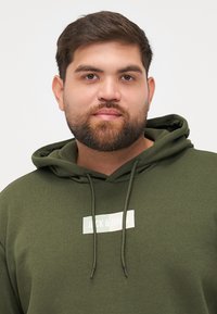 Sudadera verde oliva con capucha ajustable, que presenta un parche rectangular blanco en el centro. Tejido suave y texturizado.