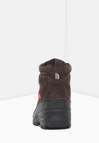 Brązowy zamszowy but hikingowy z czarną gumową podeszwą, wyściełanym kołnierzem, kontrastowym pomarańczowym logo oraz teksturowanym tyłem dla lepszej przyczepności i wsparcia.