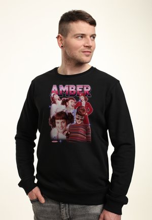 Mężczyzna noszący czarną bluzę z wieloma retro portretami kobiety i słowem „AMBER” nad nimi.