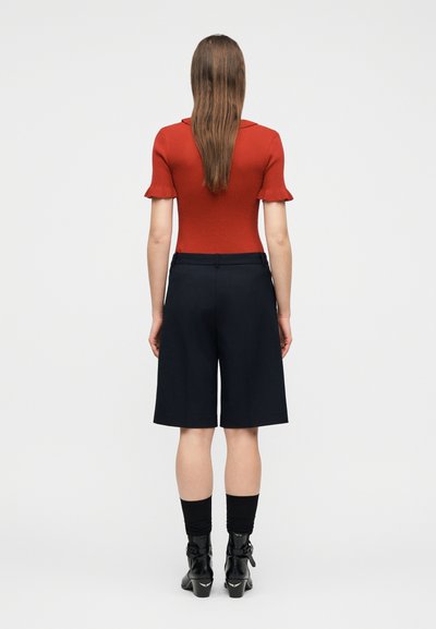 Γυναίκα με μακριά καστανά μαλλιά που φοράει εφαρμοστό κόκκινο κοντομάνικο τοπ, μαύρες σορτς μέχρι το γόνατο, μαύρες κάλτσες και μαύρα μποτάκια μέχρι τον αστράγαλο, όρθια με την πλάτη στραμμένη.