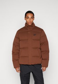 Giacca a vento marrone con colletto alto, caratterizzata da un design trapuntato e logo Nike nero sul petto. Il materiale sembra liscio e caldo.