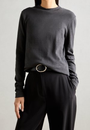 Kvinde iført en mørk, langærmet tætsiddende trøje, der er stoppet ned i sorte bukser med et sort bælte med en rund sølvspænde, hænder afslappet langs siderne.