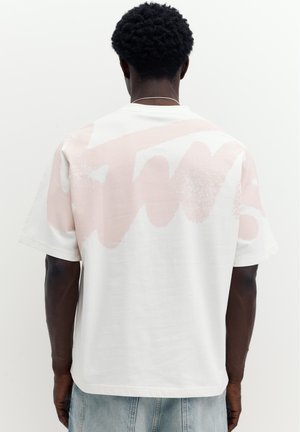 Homme aux cheveux courts et bouclés portant un t-shirt blanc avec un motif abstrait rose clair sur le haut du dos et un jean bleu clair.
