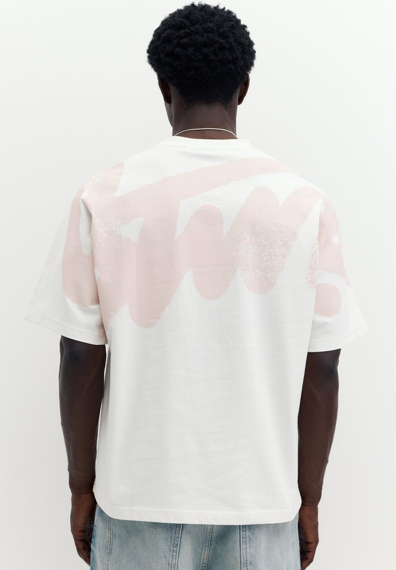 Homme aux cheveux courts et bouclés portant un t-shirt blanc avec un motif abstrait rose clair sur le haut du dos et un jean bleu clair.