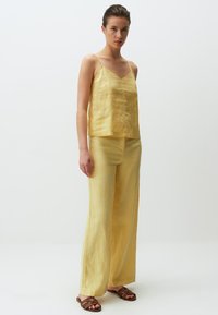 Geel linnen camisole met dunne bandjes, gecombineerd met bijpassende wide-leg broek. Lichte textuur en ontspannen pasvorm, aangevuld met bruine sandaaltjes met bandjes.