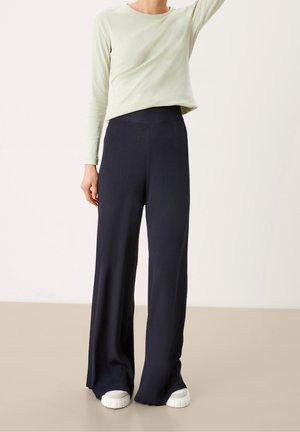 Osoba ubrana w czarne spodnie z wysokim stanem i szerokimi nogawkami oraz białe trampki, w połączeniu z jasnooliwkową bluzką z długim rękawem, stojąca na jednolitym tle.