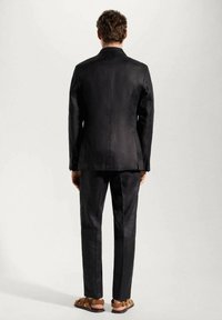 Czarny lniany garnitur o dopasowanym kroju, z pojedynczym rozporkiem z tyłu, zestawiony z brązowymi sandałami, które wyróżniają się gładką teksturą i minimalistycznym designem.