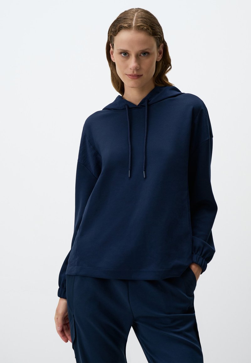 Felpa con cappuccio blu navy in tessuto morbido, caratterizzata da una vestibilità comoda, dettagli con cordoncino e polsini a coste. Abbinata a pantaloni coordinati.