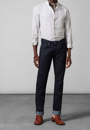 Camisa de lino blanca con botones, jeans de mezclilla oscuros con los puños enrollados, mocasines marrones y una bufanda de diseño alrededor del cuello.
