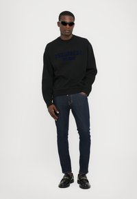 Sweat-shirt noir avec le logo bleu "DSQUARED2 MILANO", associé à un jean en denim foncé. Le modèle porte des lunettes de soleil noires et des mocassins.