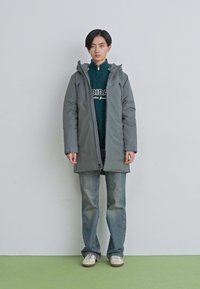 Ung person med kort hår står iført en grå hættejakke over en mørkegrøn Adidas sweatshirt, blå jeans og hvide sneakers.