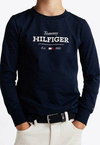Marineblaue Langarmshirt mit "Tommy HILFIGER" in weißem Text, mit Rundhalsausschnitt und taillierter Passform, kombiniert mit beigefarbenen Hosen und einem gewebten Gürtel.