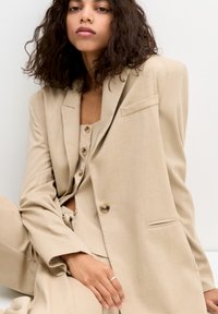 Beige striped blazer with a single button and a patch pocket, worn over a matching top. Smooth fabric with a relaxed fit.