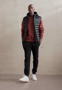 Chaleco acolchado negro sobre un sudadera color burdeos, acompañado de pantalones de chándal negros y zapatillas blancas. Texturas suaves y diseño simple.