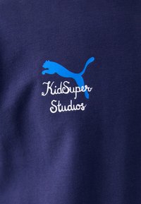 Camiseta de algodón color navy con un logo de Puma en azul claro y el texto "KidSuper Studios" en blanco, ambos ubicados en la parte superior izquierda del pecho.
