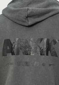 Tmavohnedá mikina s kapucňou s veľkým lesklým čiernym nápisom "ARKK" a menšími potlačenými detailmi na zadnej strane pod kapucňou.