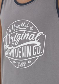 Szürke ujjatlan póló, fehér grafikával, rajta a "Original RAW DENIM CO." felirattal és további márkajelzésekkel kör alakú elrendezésben.