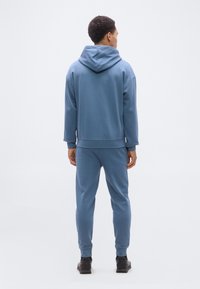 Άνδρας που στέκεται με την πλάτη, φοράει μπλε φούτερ με κουκούλα και αντίστοιχο παντελόνι φόρμας με μαύρα αθλητικά παπούτσια, σε απλό ανοιχτό φόντο.