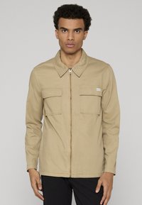Veste beige zippée avec col pointu, deux poches avant à rabat, et texture de tissu lisse. Convient pour un usage décontracté.