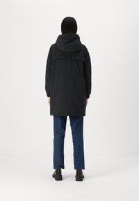 Veste à capuche noire avec taille cintrée, dotée d'un grand empeigne arrière, de poignets élastiques et d'un ourlet légèrement allongé. Associée à un jean bleu et des chaussures noires.