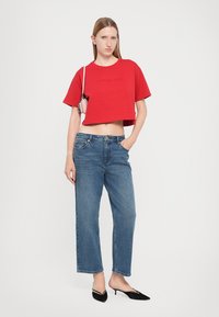 Tricou roșu scurt cu logo embosat, asortat cu blugi albaștri cu talie înaltă și croială dreaptă. Călăreți negri cu vârful ascuțit. Modelul poartă o geantă mică.
