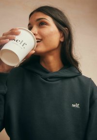 Donkergrijze hoodie met een zachte textuur, voorzien van een witte geborduurde logo "self." op de borst. Beker in de hand met een gedrukt logo.