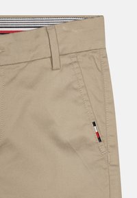 Bežové bavlněné chinos kalhoty s hladkou texturou, které mají přední kapsy a malý trojbarevný logo akcent na boku.