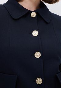 Mörkblå jacka med en strukturerad krage, utrustad med fyra texturerade guldknappar och två framfickor. Slät tyg med rena linjer.