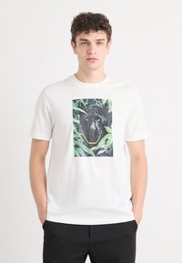 Fehér pamut póló, középen fekete párduc grafikai ábrázolással, zöld szemekkel, körülötte zöld növényzettel. Rövid ujjú, kerek nyakú.