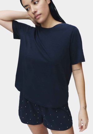 Mujer ajustándose el cabello, vestida con una camiseta azul marino oversize y pantalones cortos azul marino con pequeños puntos blancos, sobre un fondo liso.