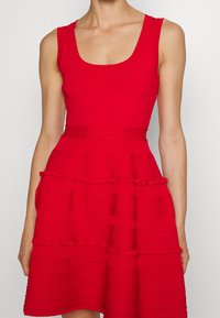 Robe rouge avec un corsage ajusté et une jupe à volants en étages. Tissu texturé avec des motifs variés et décolleté en cœur.