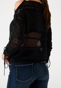 Kobieta ubrana w czarny, prześwitujący sweter z dzianiny z wzorami w zygzaki i sznureczkami, w połączeniu z ciemnoniebieskimi dżinsami.